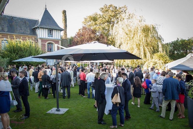 Groupe de personnes rassemblées dans un jardin pour un événement, avec des parasols et un bâtiment en arrière-plan.