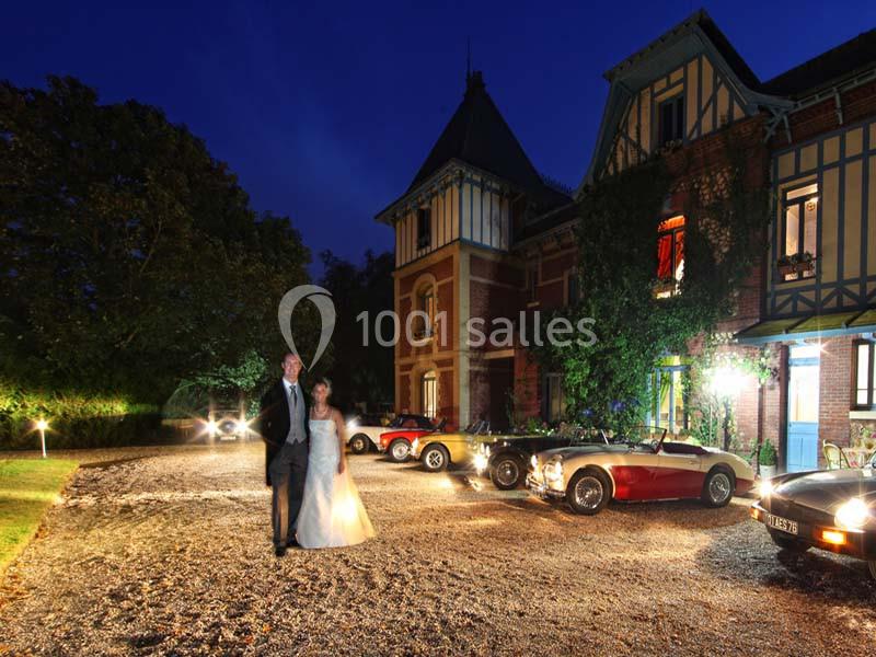 Un couple en tenue de mariage marche devant une grande maison éclairée, avec des voitures anciennes garées devant.
