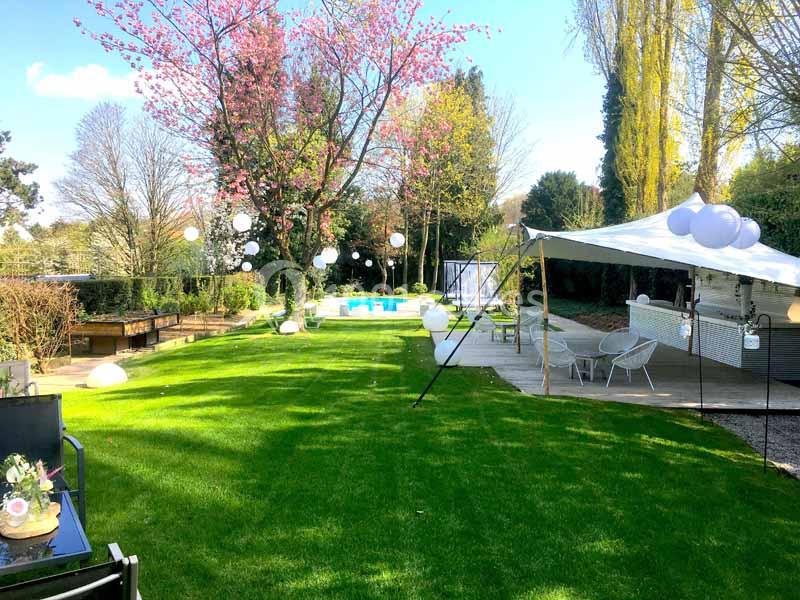 Jardin aménagé avec pelouse, arbres en fleurs, guirlandes lumineuses, piscine et espace détente sous une tente blanche.