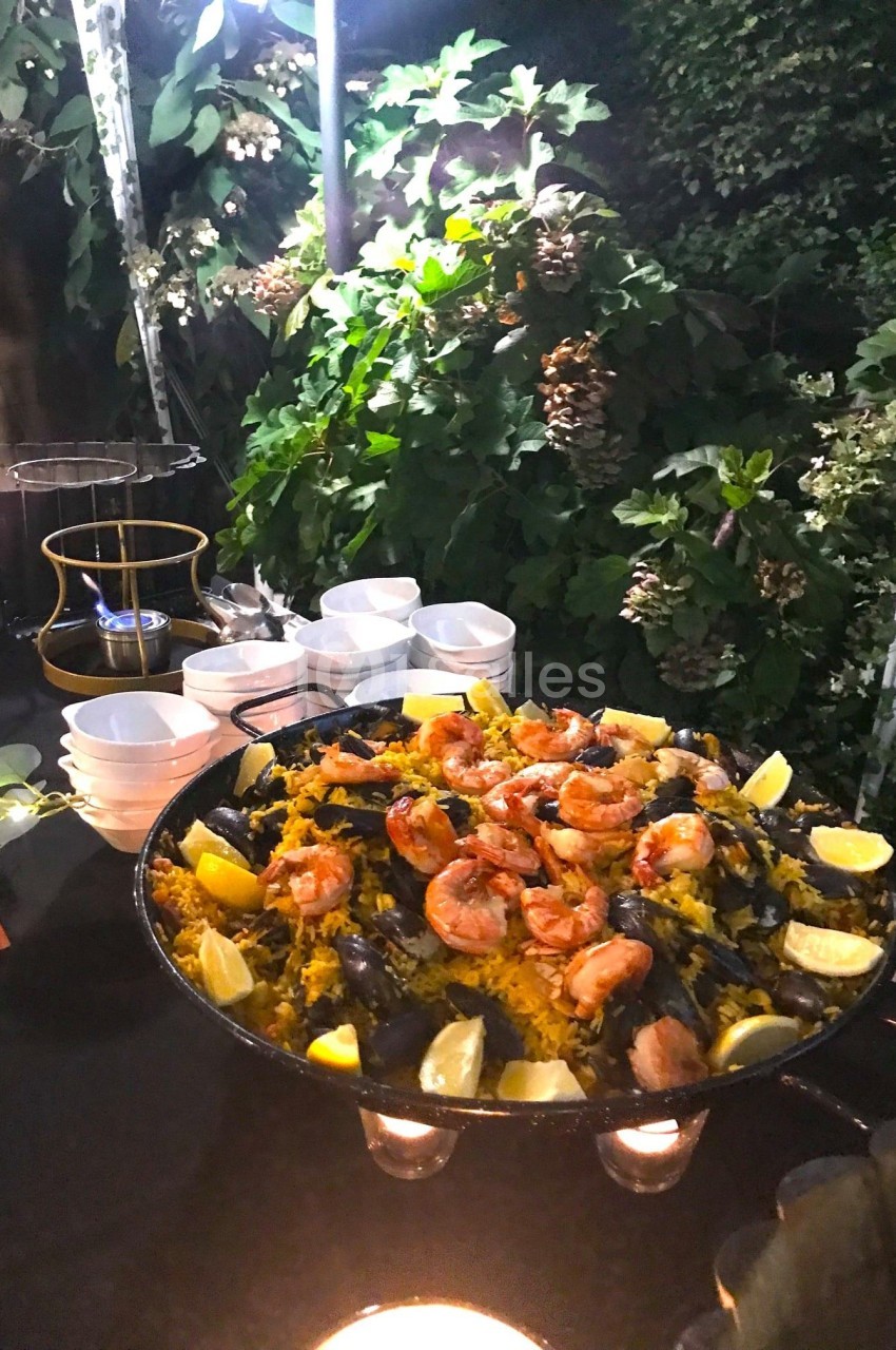 Grande poêle de paella garnie de crevettes, moules et quartiers de citron, présentée sur une table en extérieur.