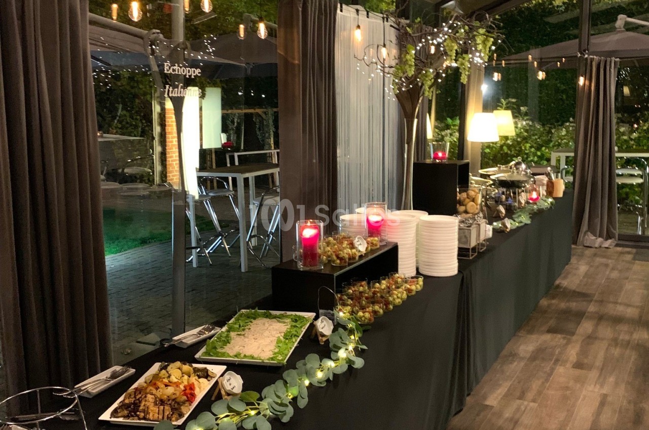 Buffet décoré avec des plats variés, bougies rouges et guirlandes lumineuses dans une salle vitrée en soirée.