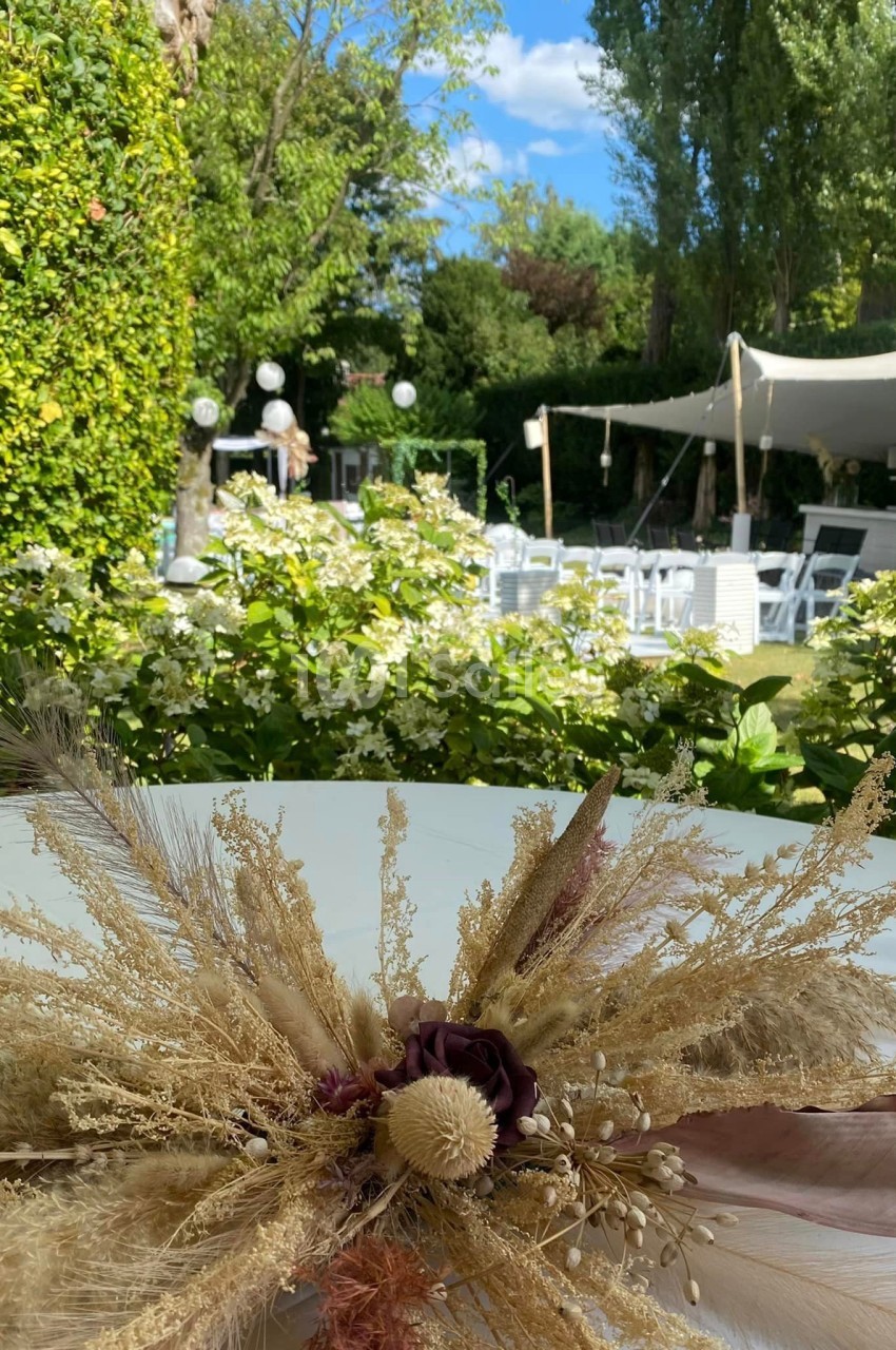 Décoration florale séchée sur une table, avec un jardin verdoyant et une tente blanche en arrière-plan.
