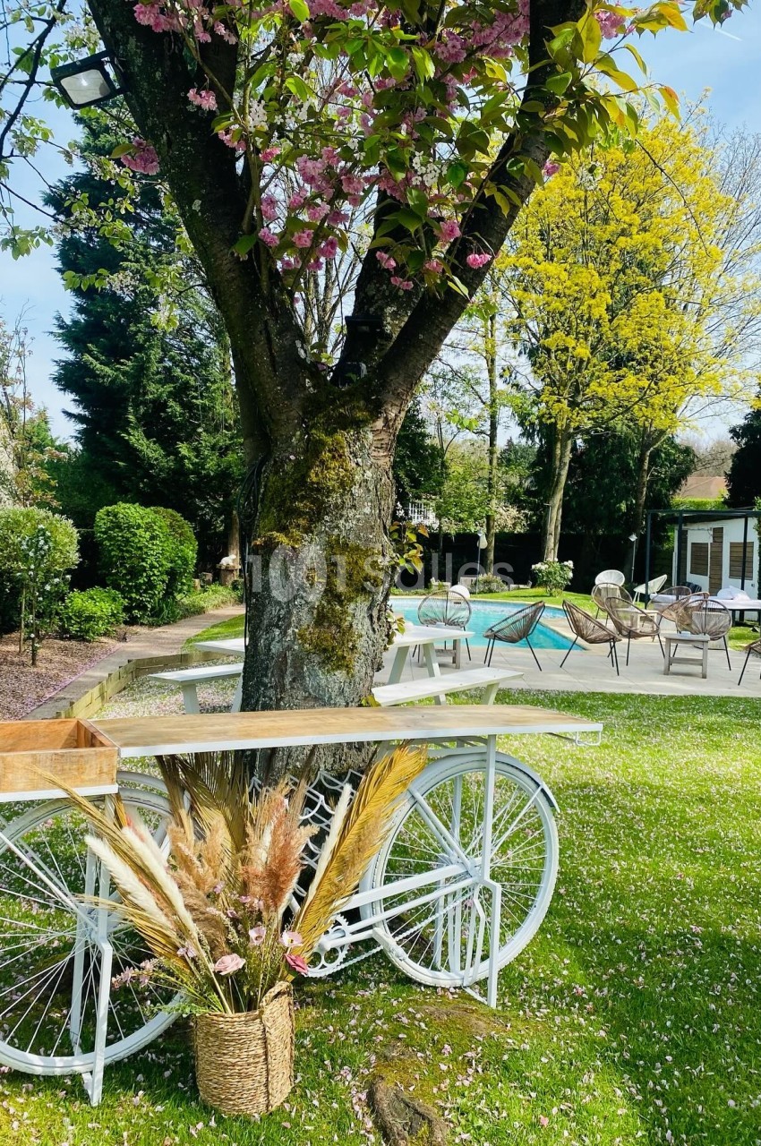Un vélo blanc décoratif avec des fleurs séchées est placé sous un arbre dans un jardin verdoyant près d'une piscine.