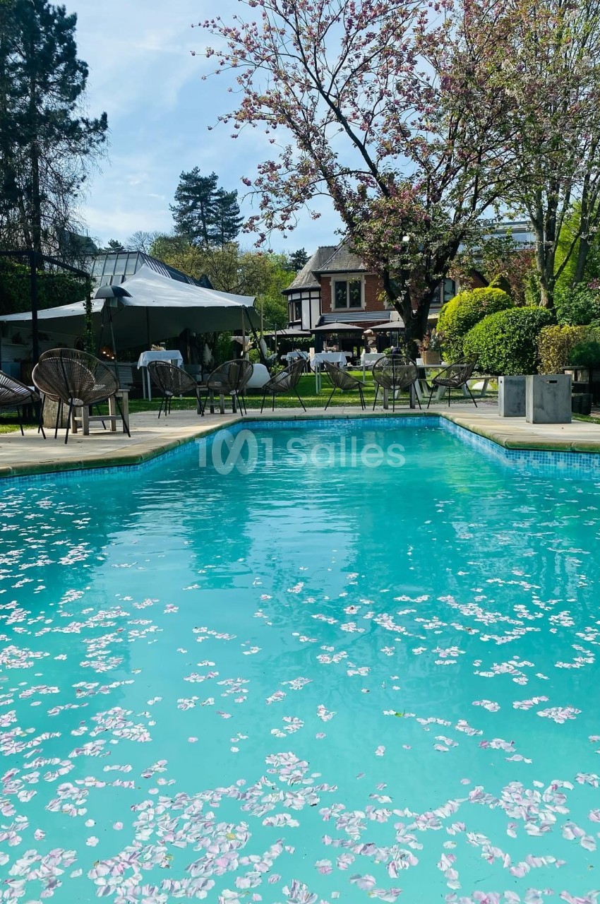 Piscine extérieure avec pétales de fleurs flottant à la surface, entourée d'arbres et d'une maison en arrière-plan.