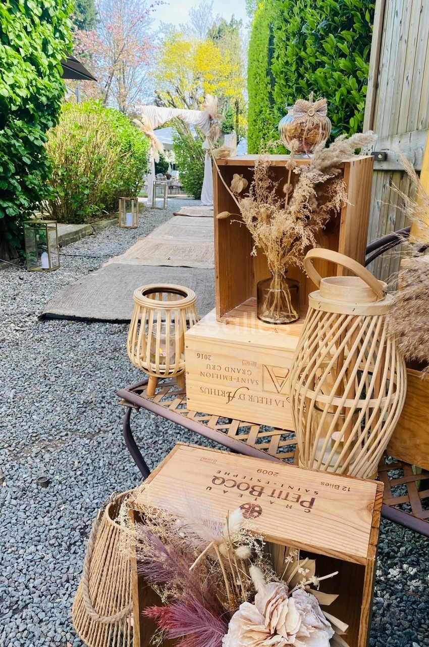 Décoration champêtre avec caisses en bois, lanternes en osier et fleurs séchées sur un chemin de gravier bordé de verdure.