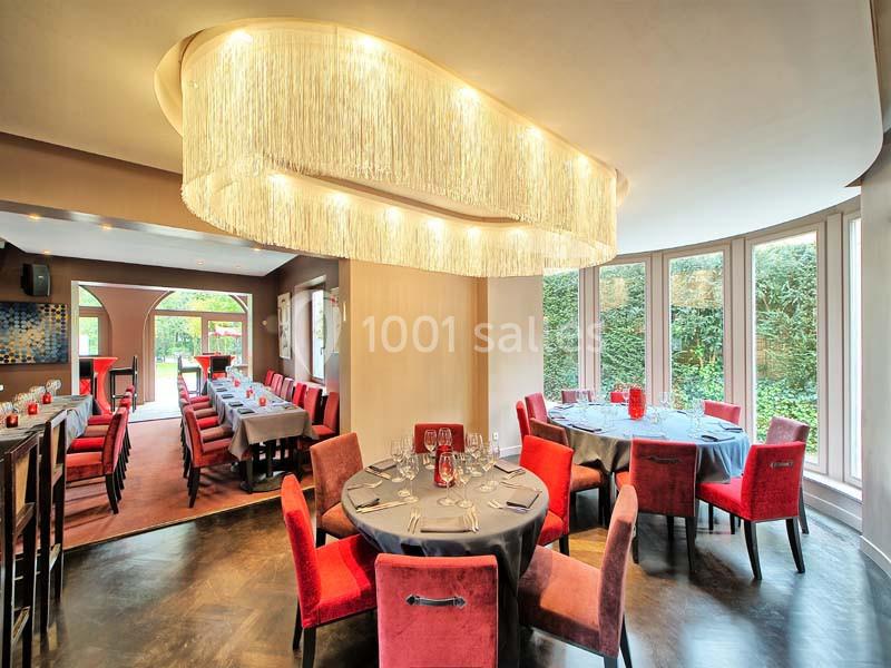 Salle de restaurant lumineuse avec tables dressées, chaises rouges et grand lustre moderne, donnant sur un jardin.