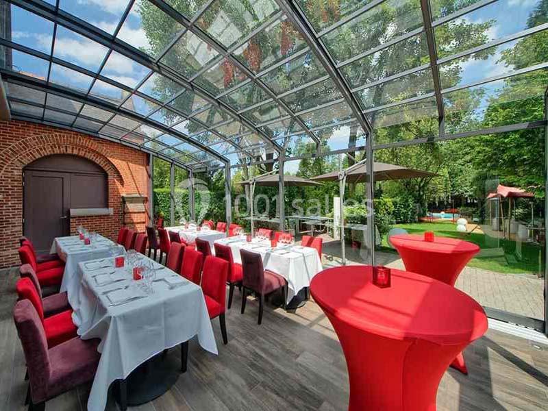 Salle lumineuse avec tables dressées sous une verrière, chaises rouges et vue sur un jardin verdoyant.
