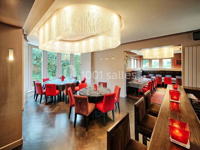 Salle de restaurant lumineuse avec tables rondes dressées, chaises rouges et grandes suspensions modernes.