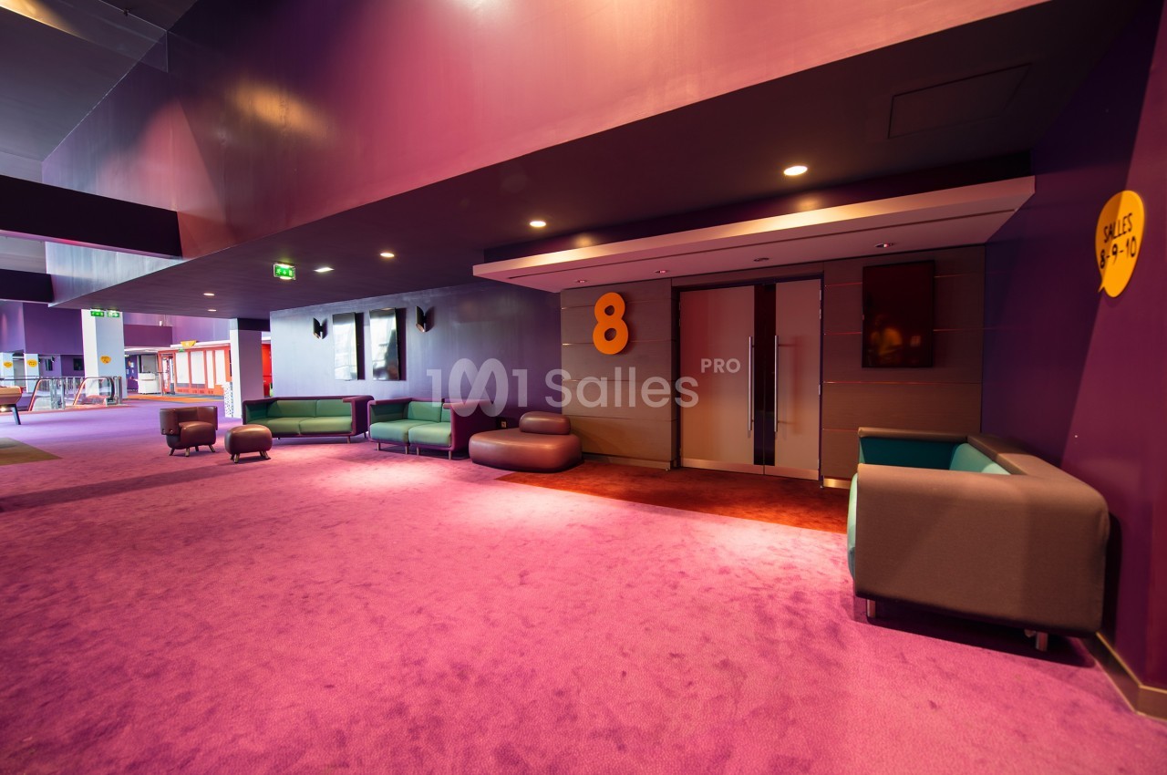 Couloir d'un cinéma avec moquette violette, canapés modernes, et porte numérotée ’8’ menant à une salle.