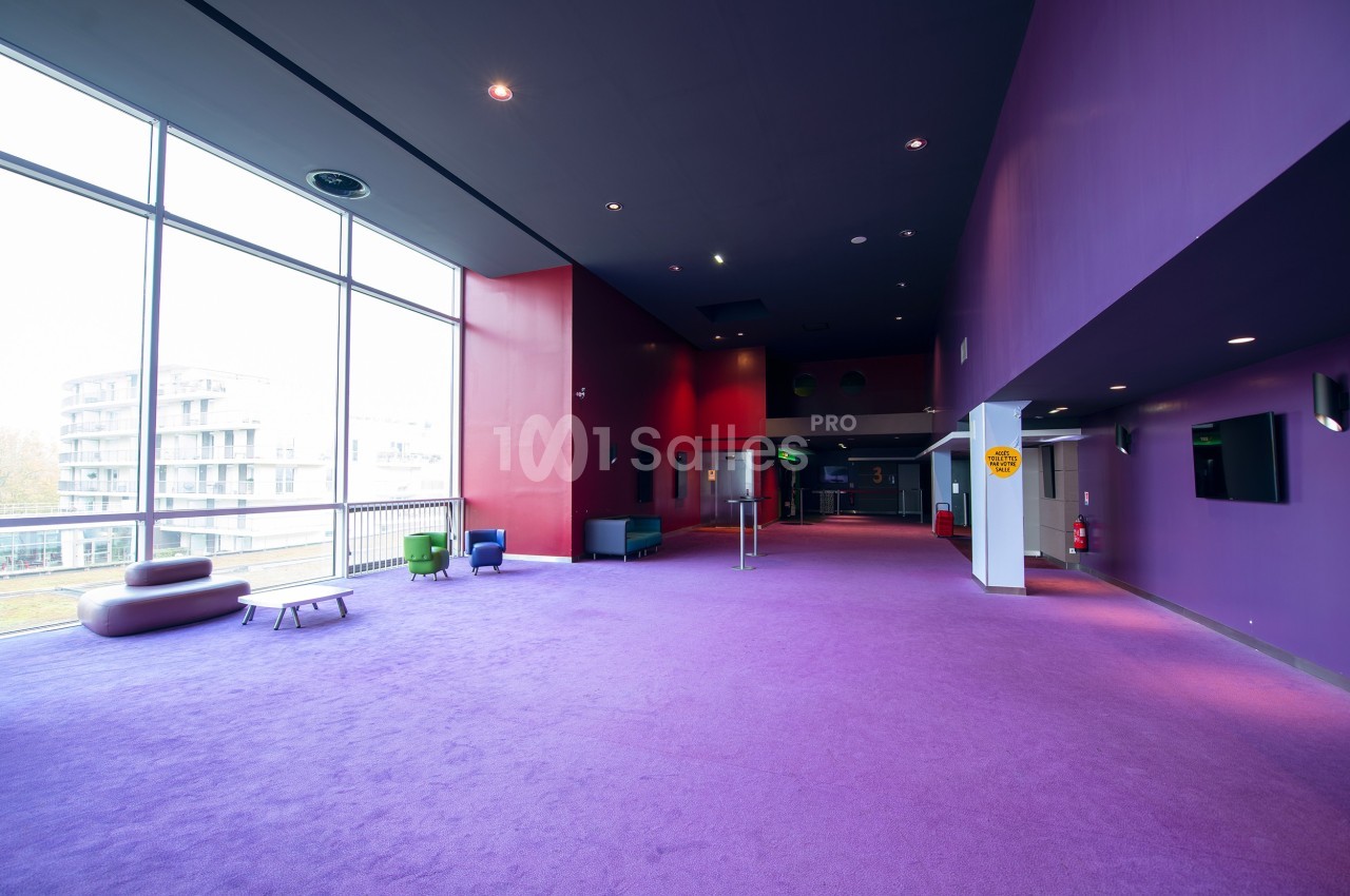 Hall spacieux avec moquette violette, murs colorés et grandes baies vitrées laissant entrer la lumière naturelle.