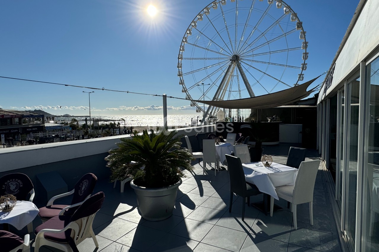 Terrasse ensoleillée avec des tables dressées, vue sur une grande roue et la mer en arrière-plan.