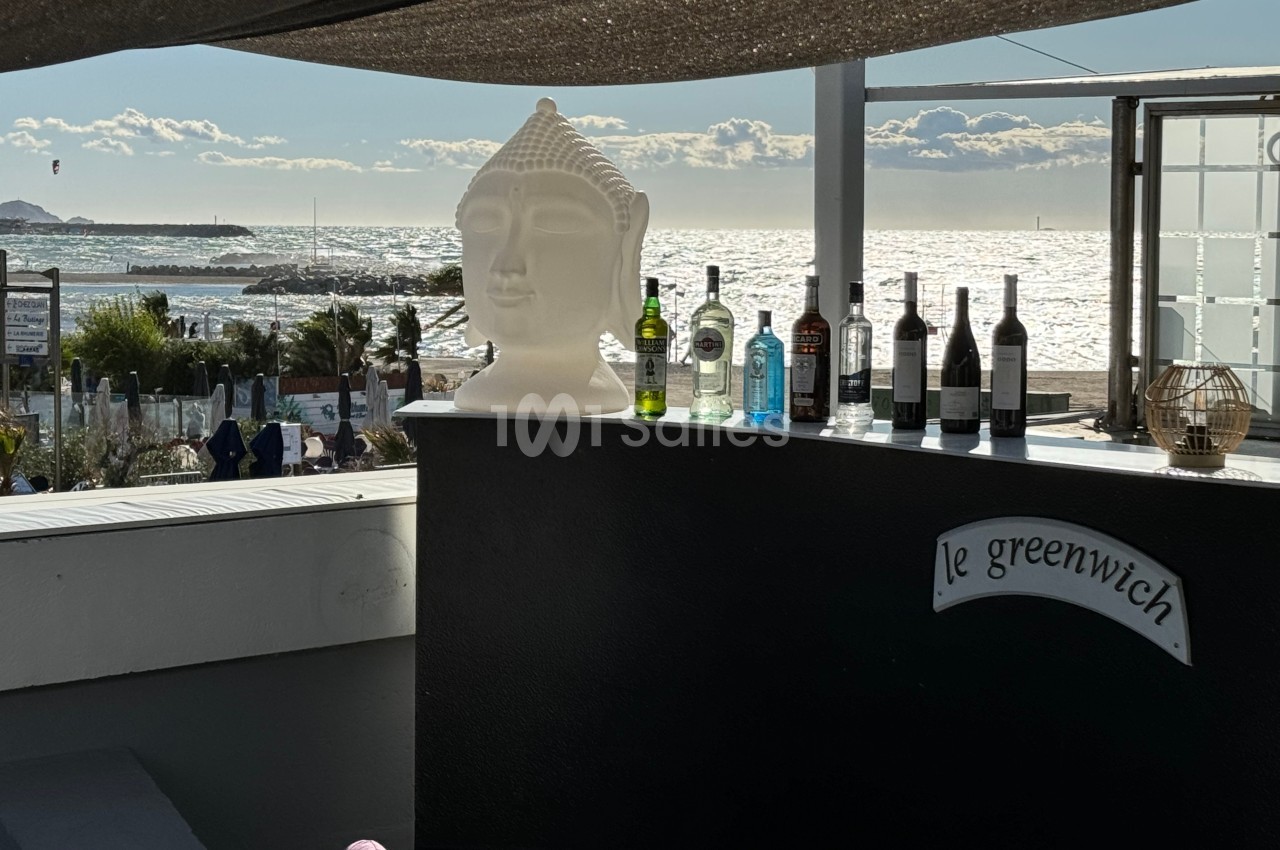 Statue de Bouddha et bouteilles alignées sur un comptoir en terrasse avec vue sur la mer et le ciel ensoleillé.