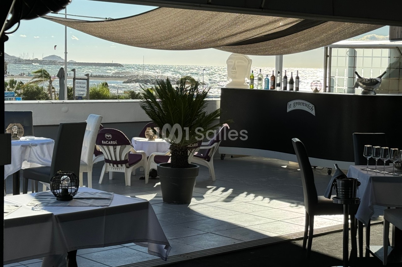Terrasse d'un restaurant avec vue sur la mer, tables dressées, palmier en pot et comptoir avec bouteilles exposées.