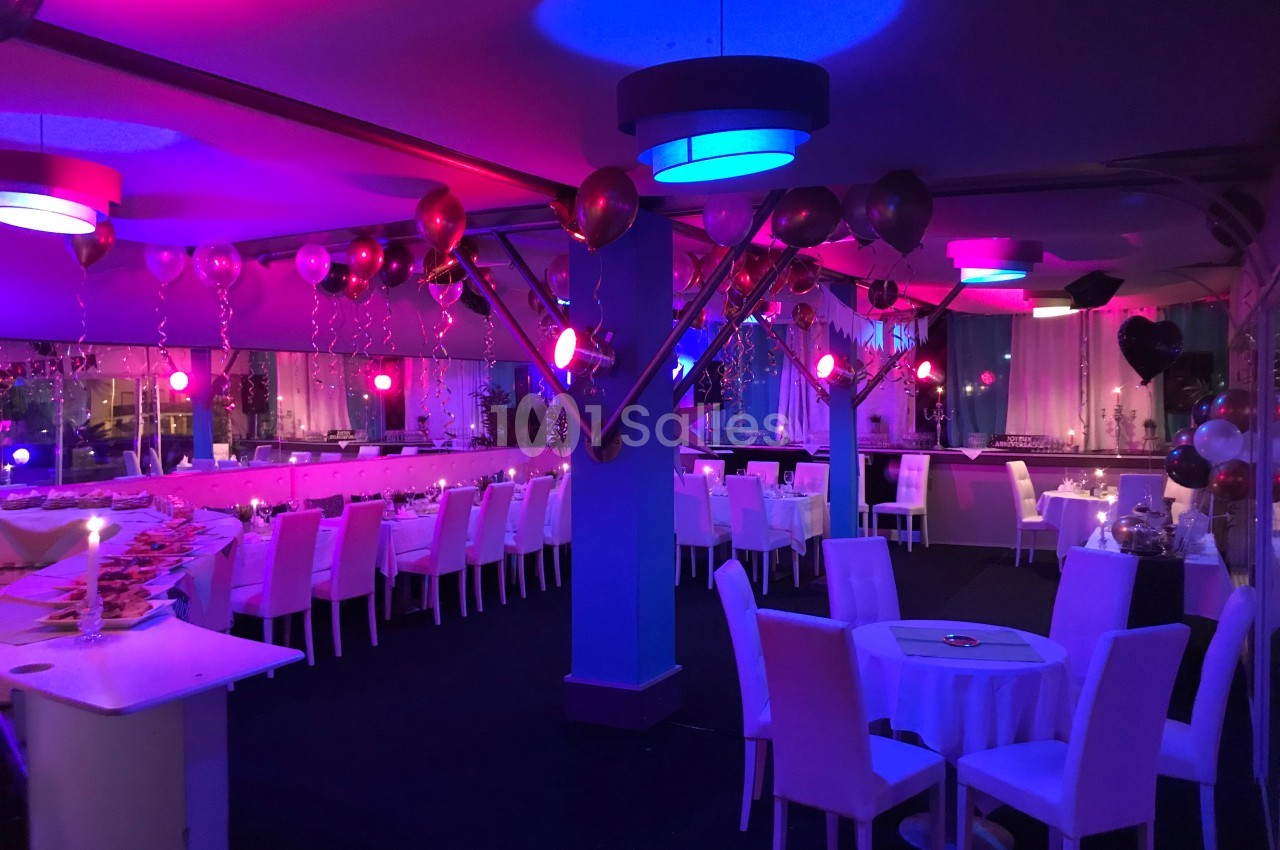 Salle décorée pour un événement avec des ballons, des tables dressées et un éclairage coloré rose et bleu.