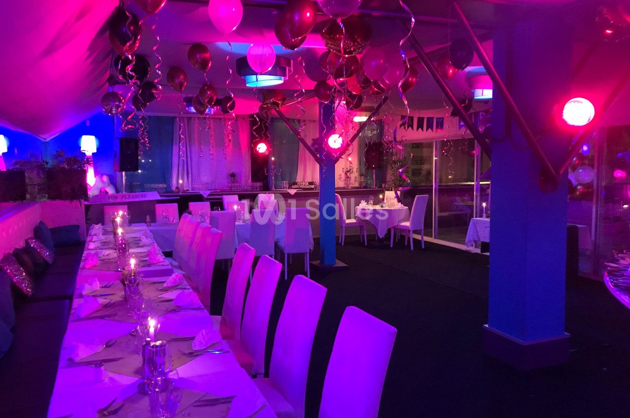 Salle décorée pour un événement festif avec tables dressées, ballons suspendus et éclairage coloré.