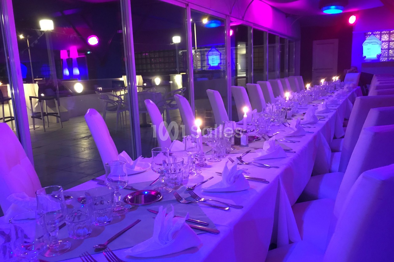 Table dressée avec nappes blanches, couverts et verres, dans une salle éclairée par des lumières violettes et bleues.