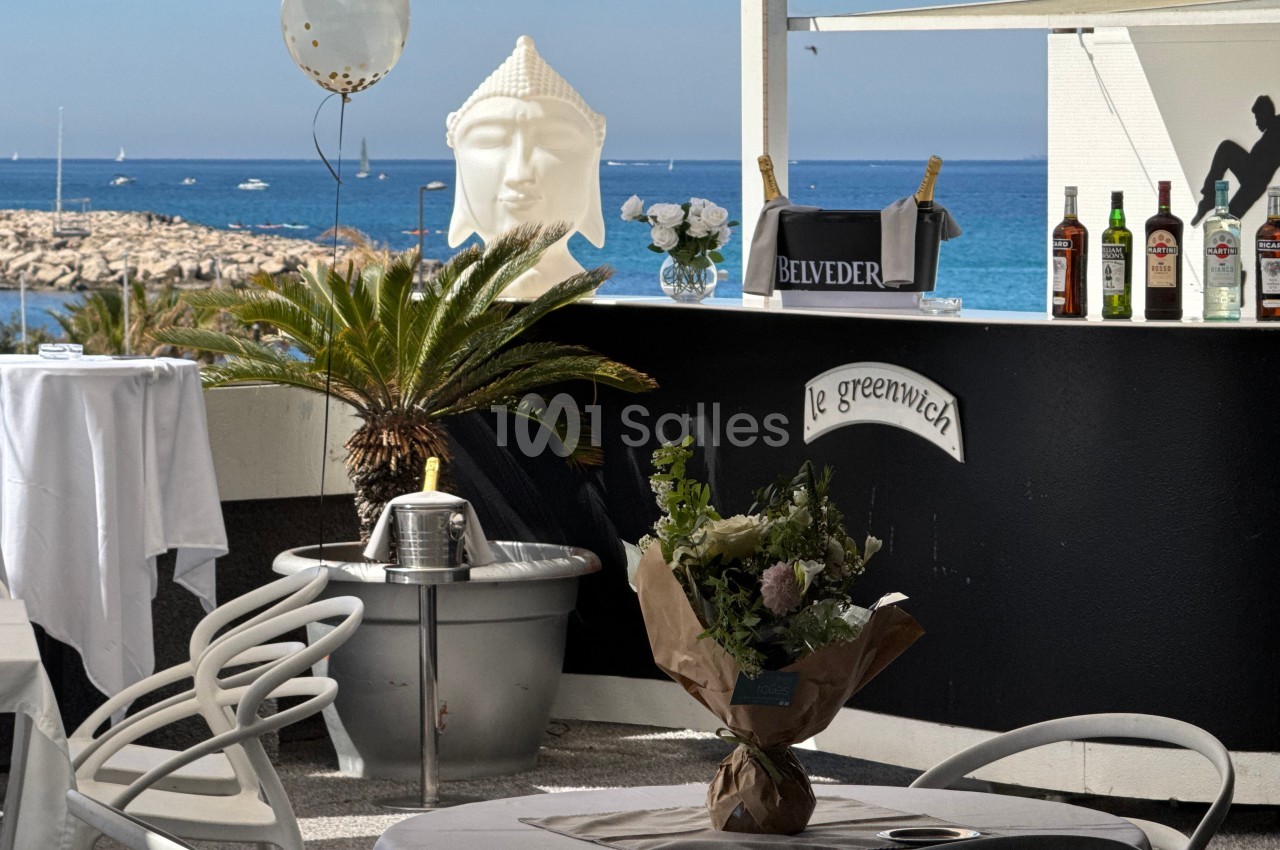 Bar extérieur avec vue sur la mer, décoré de plantes, statues et bouteilles, table dressée au premier plan.