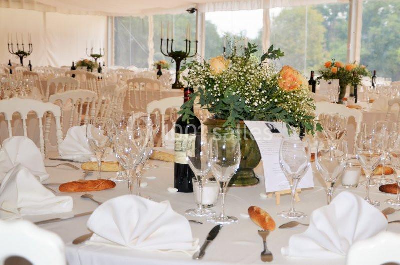 Table dressée pour un événement avec nappes blanches, verres, fleurs, bougies, pain et une bouteille de vin.