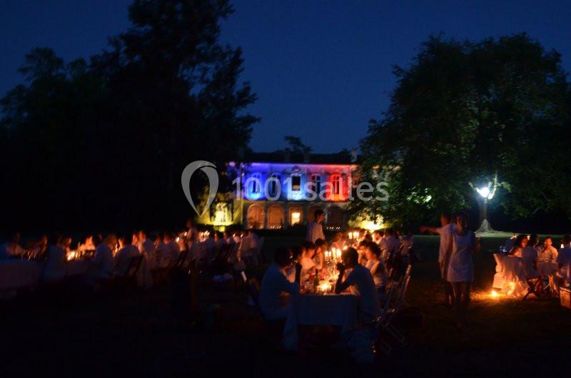 Des convives vêtus de blanc dînent à l'extérieur, éclairés par des bougies, devant un bâtiment illuminé aux couleurs bleu…