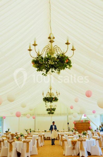 Intérieur d'une tente de réception décorée avec des lustres ornés de verdure, des tables dressées et des lanternes…