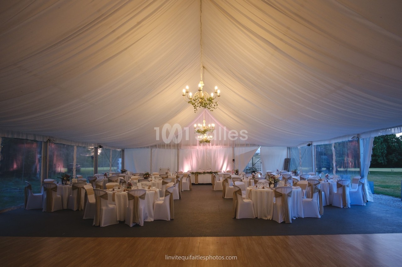Intérieur d'une tente de réception élégamment décorée avec des tables rondes, des chaises blanches et des lustres suspendus.