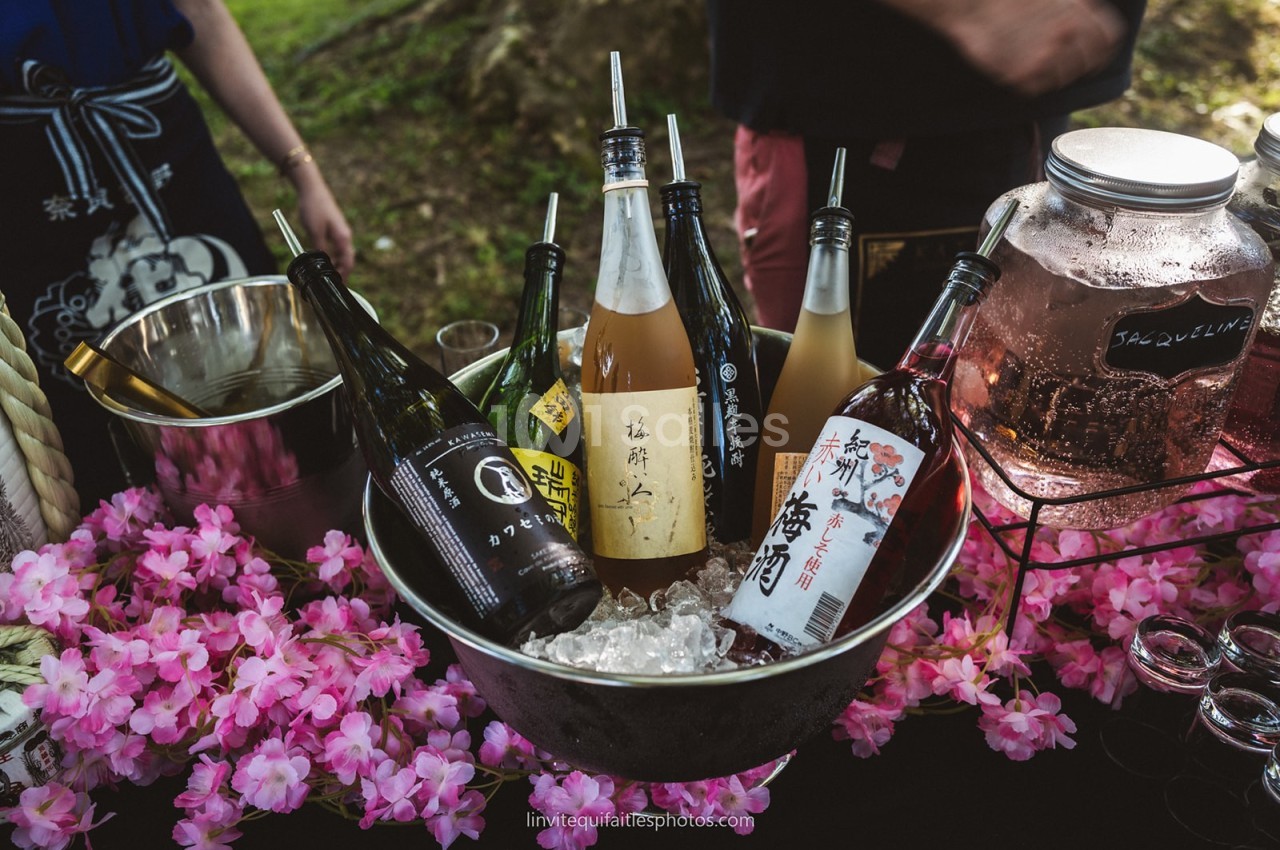 Bouteille de saké et autres boissons disposées sur de la glace dans un seau, entourées de fleurs roses décoratives.