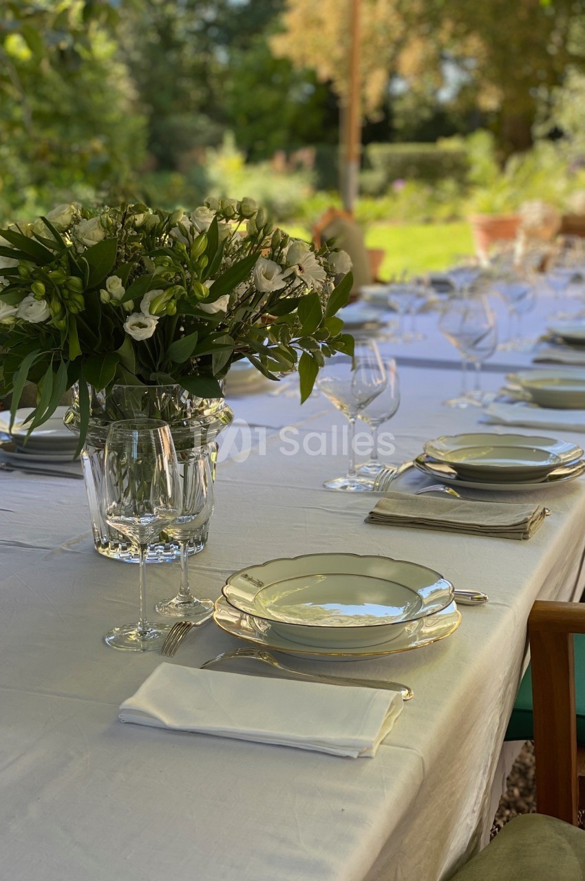 Table dressée en extérieur avec nappes blanches, vaisselle élégante, verres et bouquet de fleurs au centre.