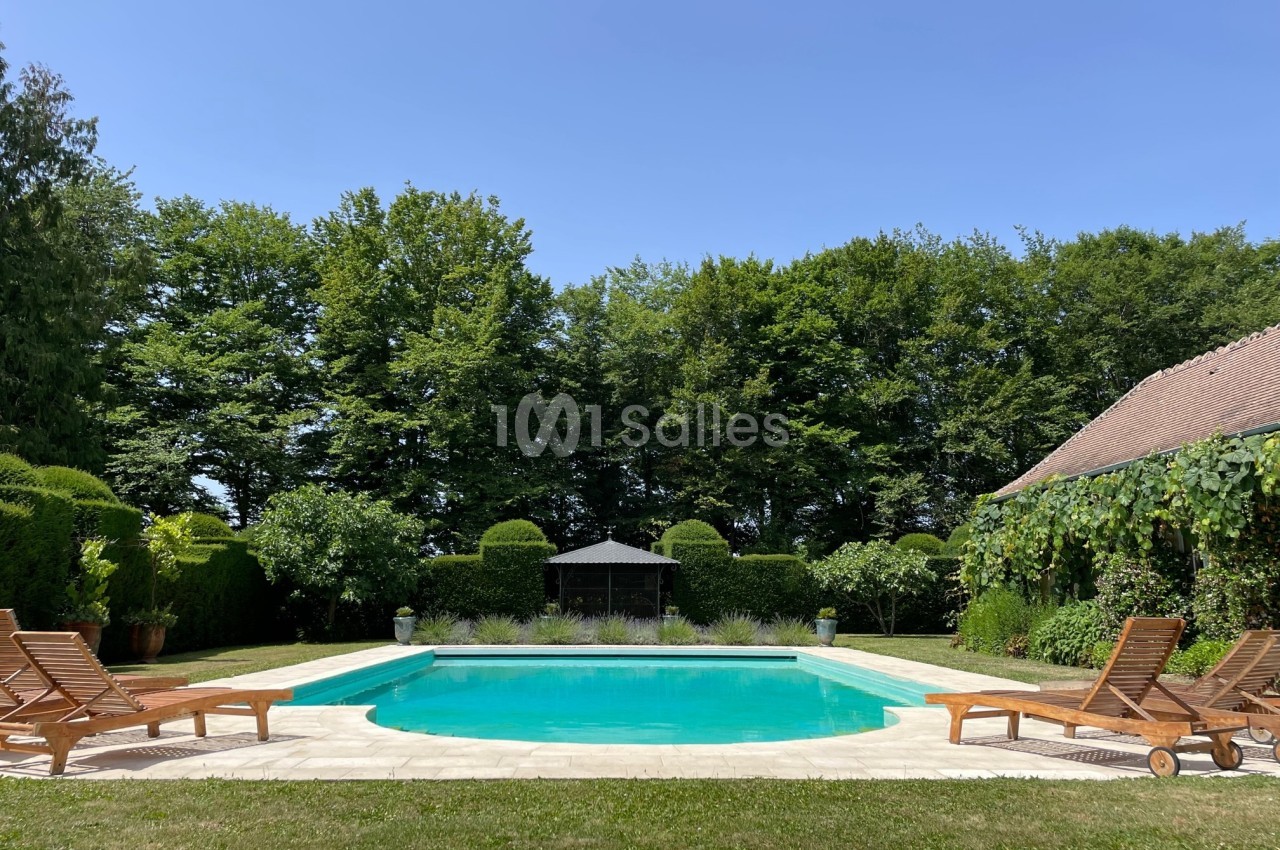 Piscine extérieure entourée de chaises longues en bois, pelouse, arbres et cabanon sous un ciel dégagé.