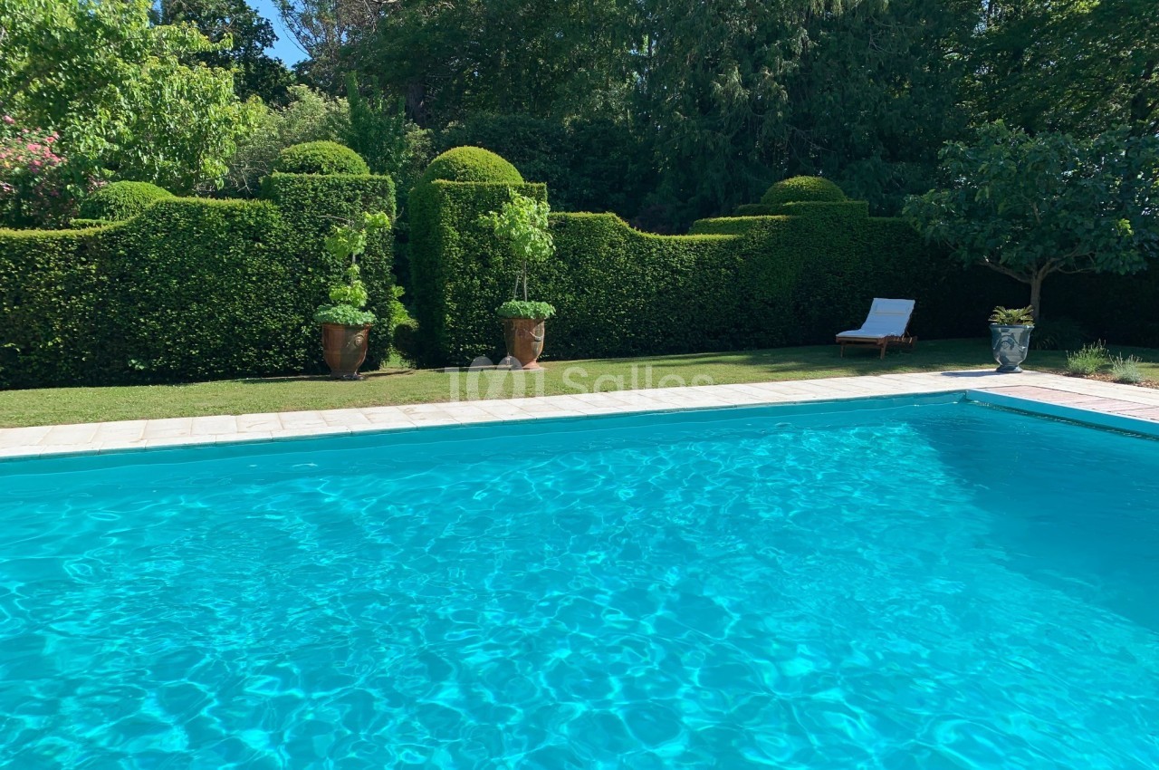 Piscine extérieure entourée de haies taillées, avec un transat blanc et des pots de plantes en arrière-plan.