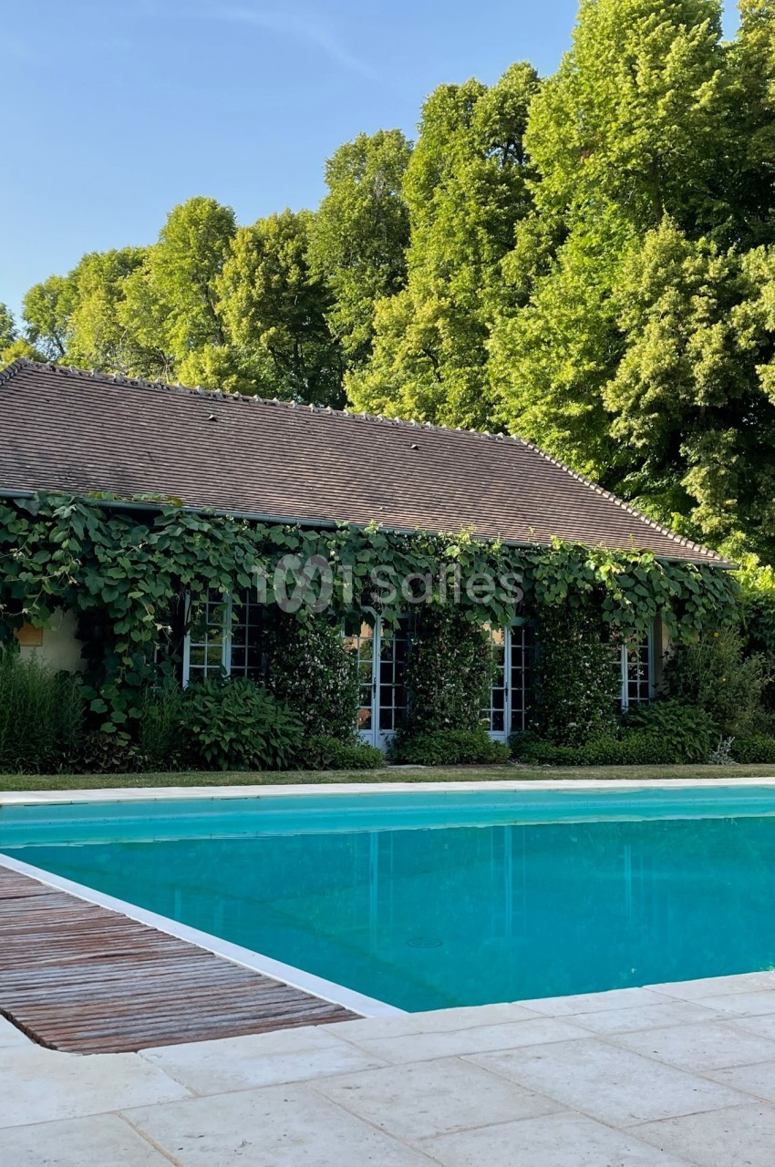 Maison entourée de verdure avec une piscine au premier plan et des arbres en arrière-plan sous un ciel dégagé.
