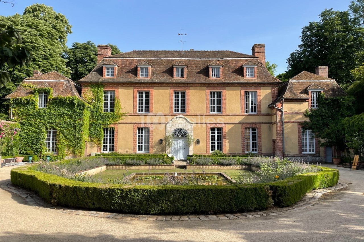 Façade d'un manoir ancien entouré de verdure, avec une fontaine centrale et un jardin bien entretenu.