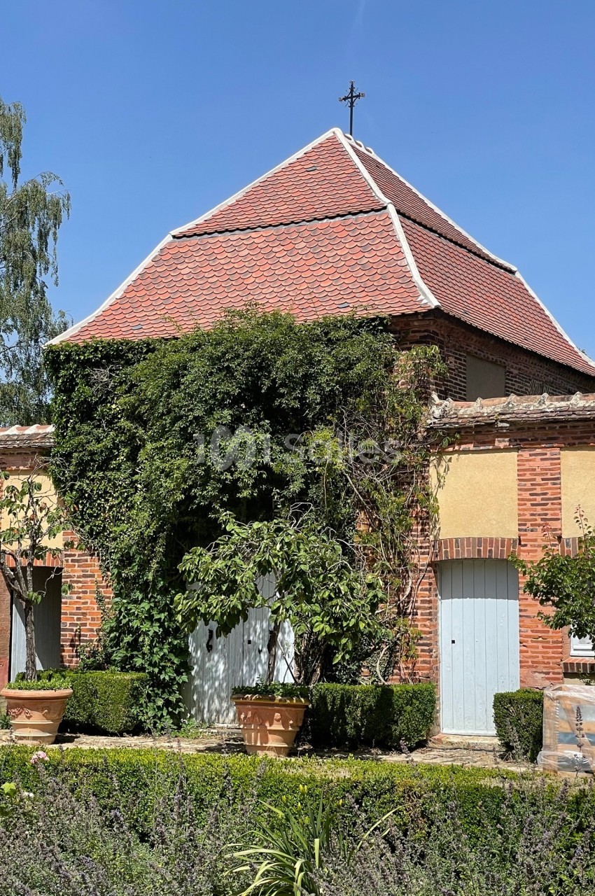 Bâtiment en briques avec toit en tuiles rouges, entouré de plantes grimpantes et d'arbustes taillés.