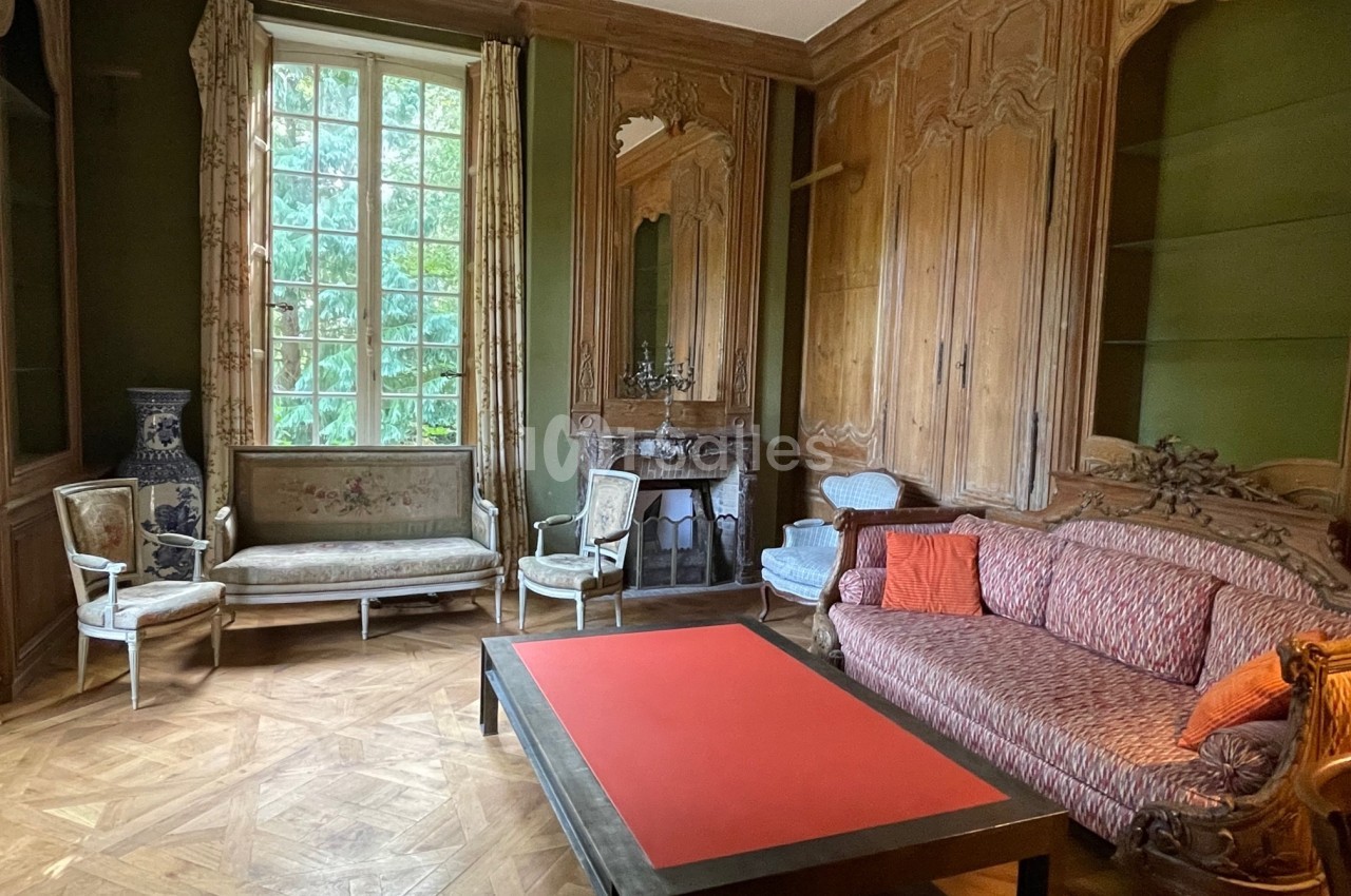 Salon ancien avec boiseries sculptées, cheminée, meubles d'époque et grande fenêtre donnant sur un jardin verdoyant.