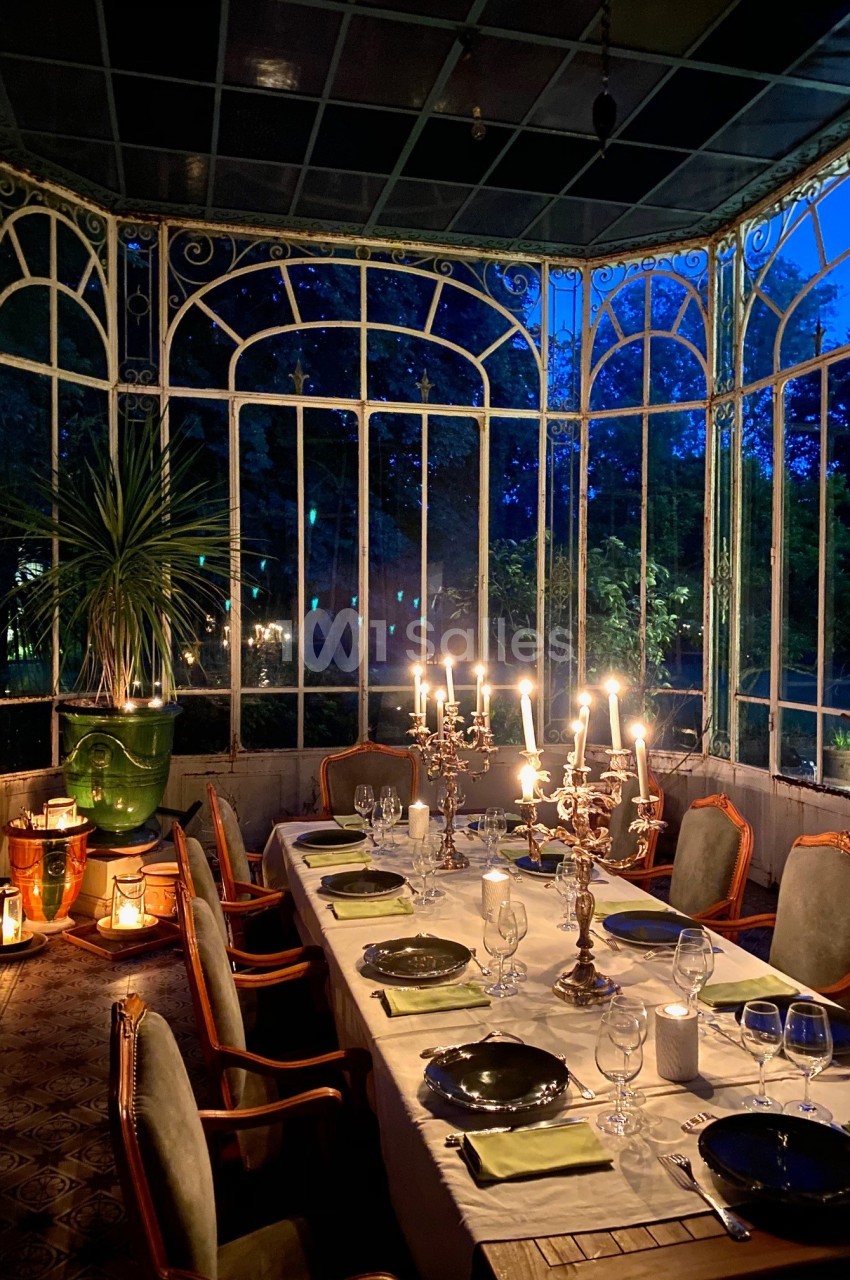 Table dressée avec chandeliers allumés dans une verrière élégante, entourée de végétation visible à travers les vitres.