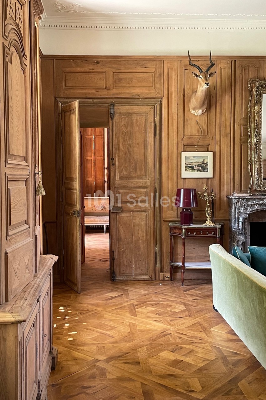 Intérieur d'une pièce avec boiseries murales, parquet, cheminée, canapé vert et tête d'antilope accrochée au mur.