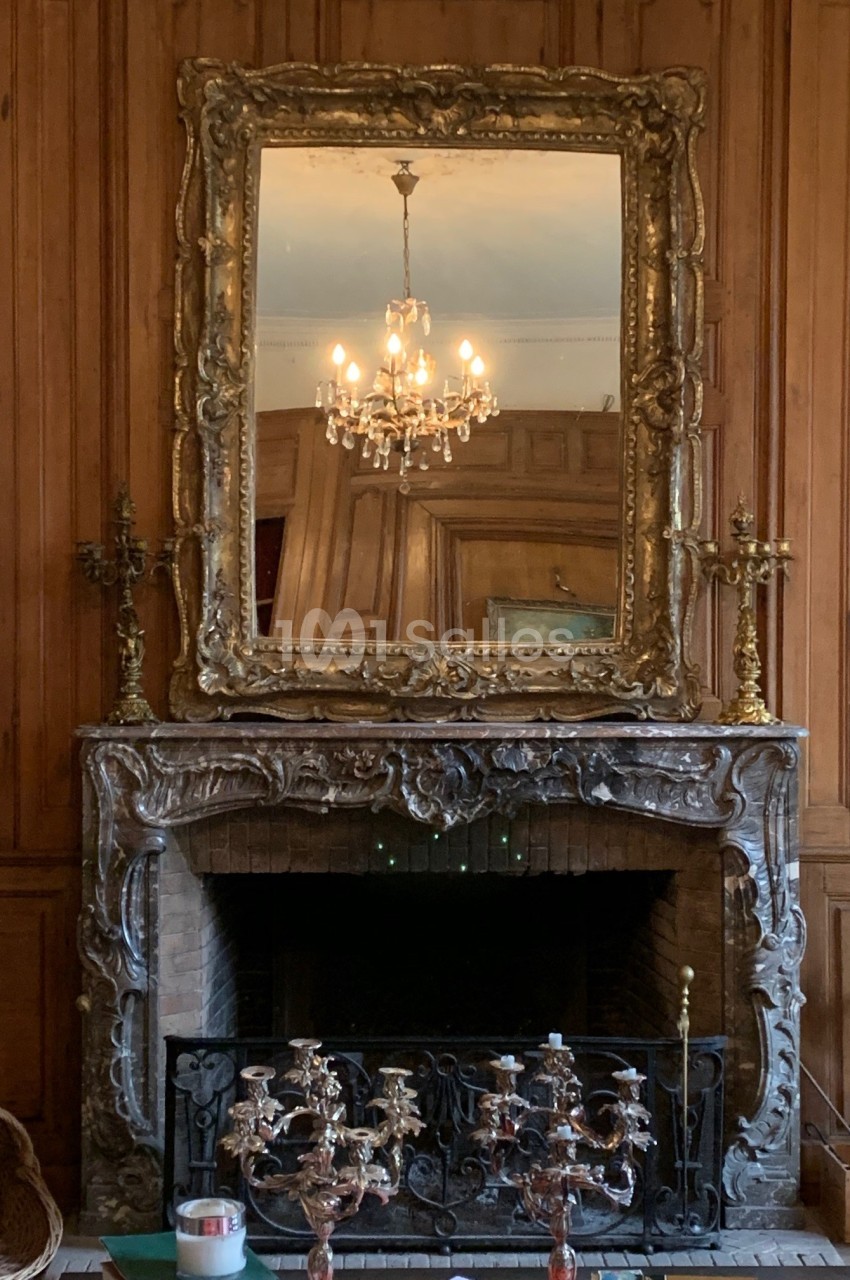 Miroir doré au-dessus d'une cheminée en marbre sculpté, entouré de boiseries et éclairé par un lustre.