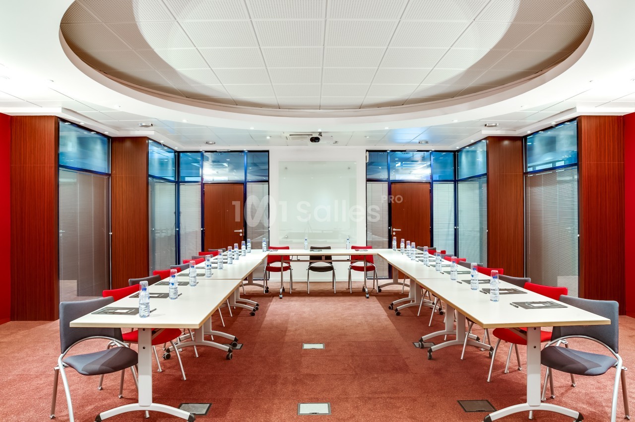 Salle de réunion moderne avec tables disposées en U, chaises rouges et grises, bouteilles d'eau et éclairage lumineux.