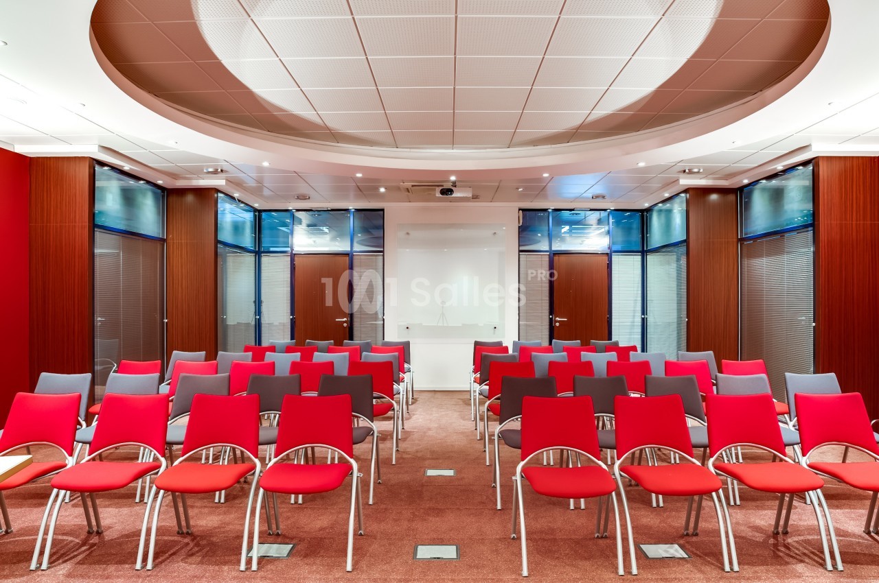 Salle de conférence moderne avec rangées de chaises rouges et grises face à un tableau blanc.