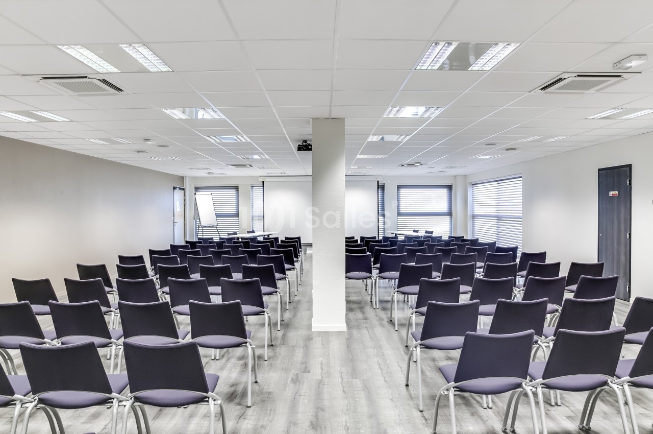 Salle de conférence moderne avec rangées de chaises vides, éclairée par des fenêtres et des lumières au plafond.