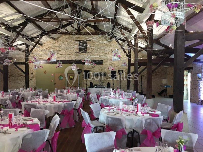 Salle de réception décorée avec des tables rondes, nappes blanches, nœuds roses et guirlandes suspendues.