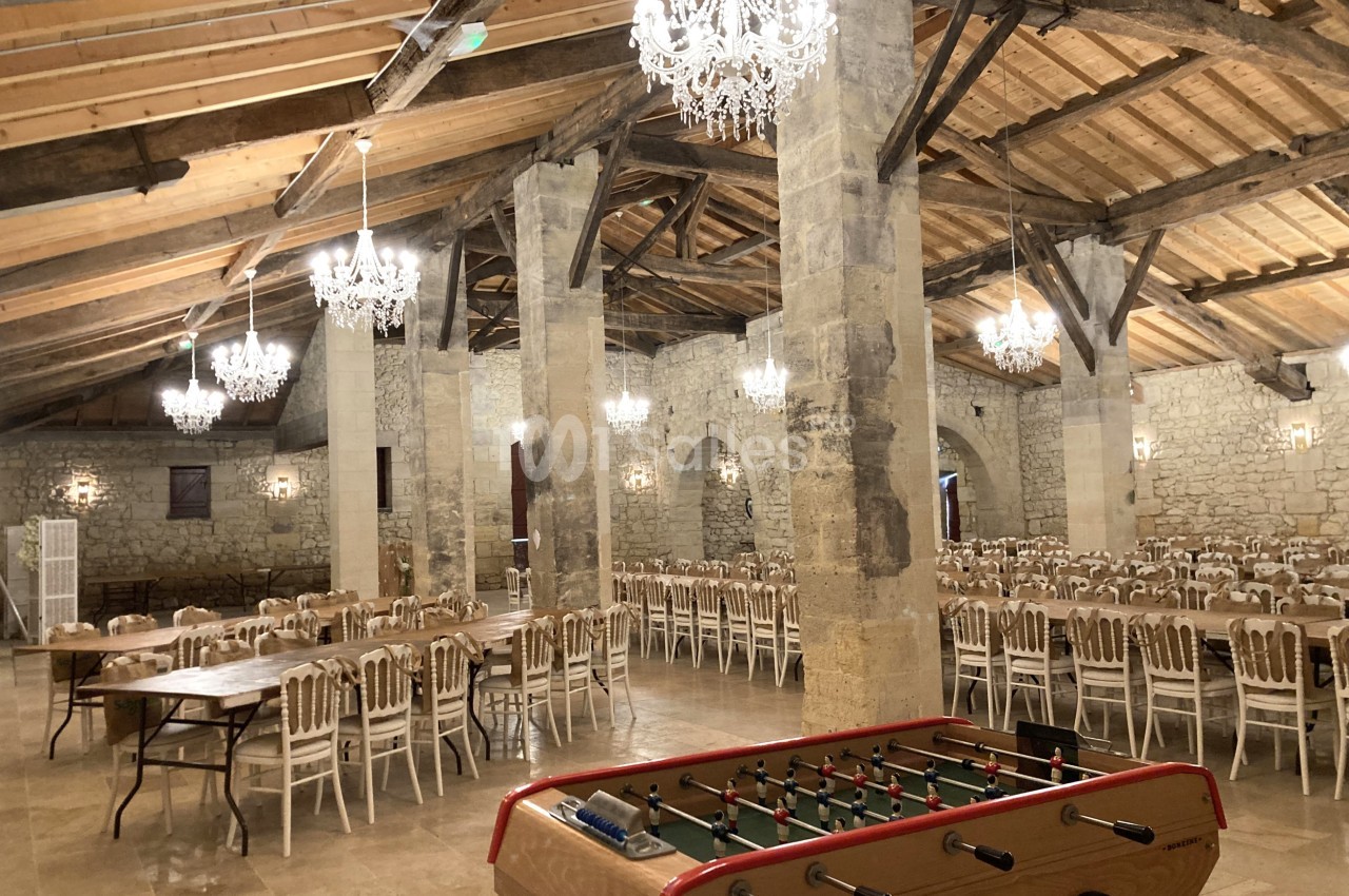 Salle en pierre avec poutres apparentes, lustres suspendus, tables dressées et un baby-foot au premier plan.