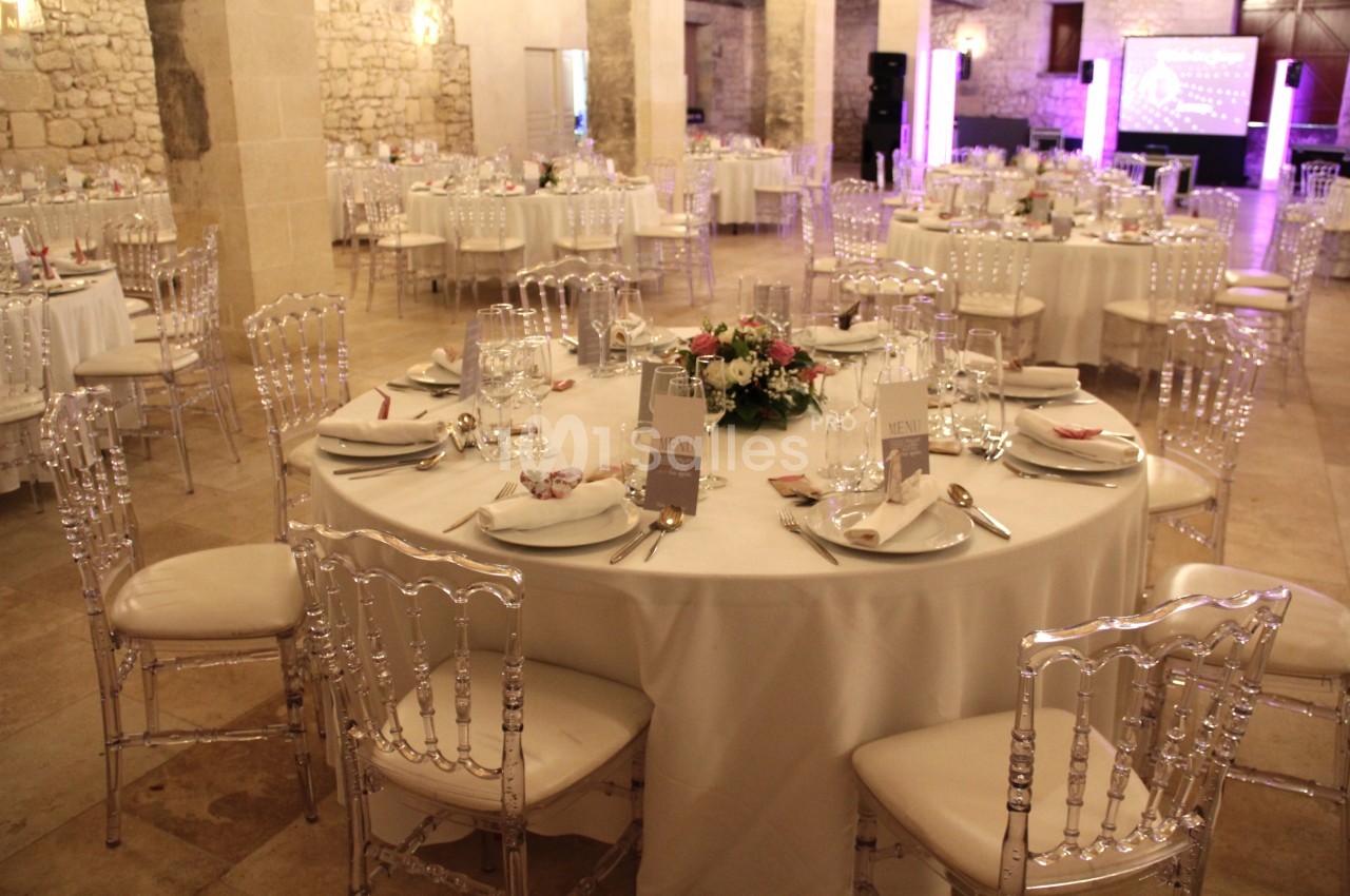 Salle de réception avec tables rondes dressées, nappes blanches, chaises transparentes et décoration florale centrale.