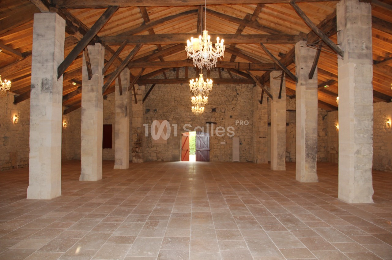 Salle spacieuse avec murs en pierre, poutres en bois apparentes et lustres suspendus, éclairée par une porte ouverte.