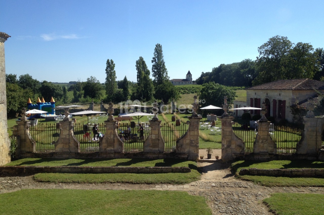 Vue d'un jardin avec pelouse, tables, parasols et une structure gonflable, entouré de bâtiments anciens et d'arbres.