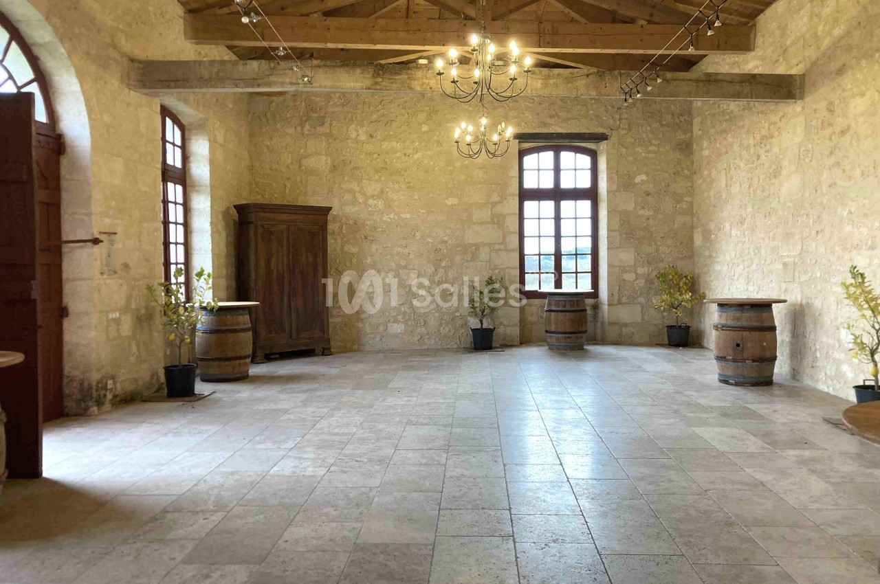 Salle en pierre avec plafond en poutres apparentes, fenêtres lumineuses, tonneaux en bois et plantes en pots.