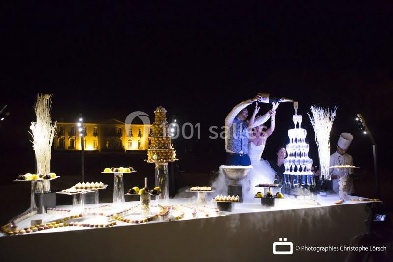 Un couple verse du champagne sur une fontaine illuminée, entouré de desserts, devant un bâtiment éclairé la nuit.