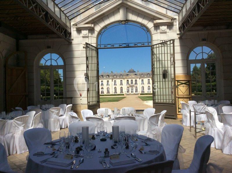 Salle de réception lumineuse avec tables rondes dressées, vue sur un jardin et un bâtiment historique en arrière-plan.