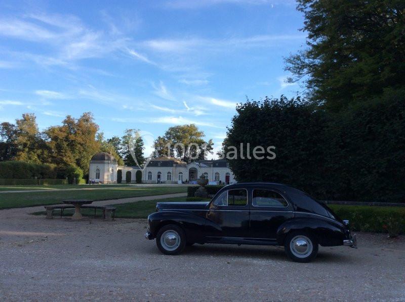 Voiture ancienne noire stationnée devant un bâtiment blanc entouré de jardins et d'arbres sous un ciel bleu.