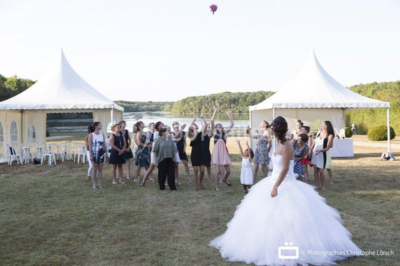 Une mariée lance son bouquet à un groupe de femmes rassemblées sur une pelouse devant des tentes blanches.