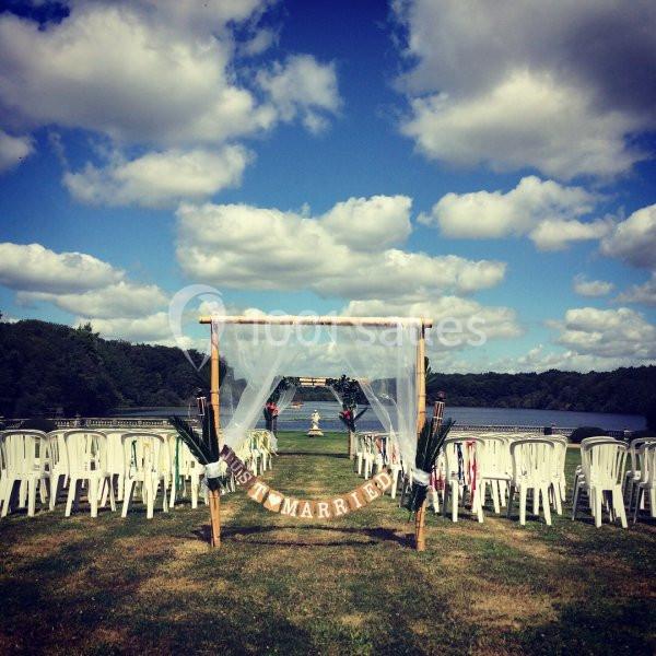 Arche de mariage décorée avec des voilages blancs, entourée de chaises blanches, face à un lac sous un ciel bleu.